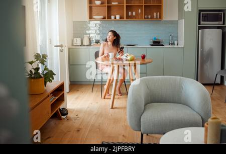 Femme au téléphone pensant à l'idée de petit déjeuner, en-cas sain tout en buvant du café le matin dans la cuisine élégante. Décontracté et seule fille à la recherche Banque D'Images