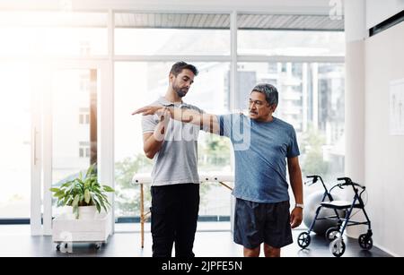 Étirez-vous aussi loin que possible. Un jeune kinésithérapeute masculin aidant un patient mâle mature avec des exercices de mouvement dans une clinique. Banque D'Images