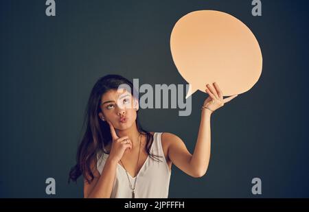 Laissez-moi réfléchir un instant. Photo en studio d'une jeune femme insouciante tenant une bulle de parole tout en contemplant et en se tenant debout sur un fond sombre Banque D'Images