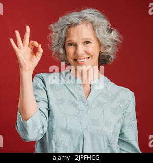 Bon, parfait et d'accord avec un geste de main de femme senior ou signe dans le soutien ou le soutien positif dans le studio sur un fond rouge. Portrait d'un Banque D'Images