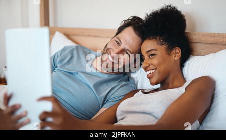 Couple interracial heureux, détendu et insouciant sur une tablette numérique souriant et prenant un selfie dans le lit. Mari et femme se réveillant et lisant les médias sociaux Banque D'Images