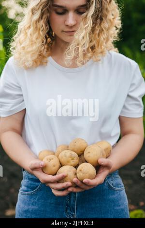 Femme maurique dans un T-shirt blanc tenant de jeunes pommes de terre maison dans ses mains. Nourriture biologique, légumes, agriculture, gros plan. Banque D'Images