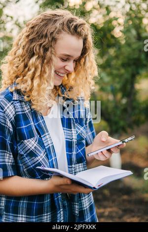 Femme souriante aux cheveux bouclés utilisant le téléphone portable et étudiant le développement des pruniers. Une agriculture intelligente. Technologie agricole et biologique Banque D'Images