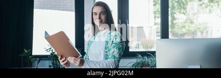 Jeune couturier tenant la chemise en papier et regardant l'appareil photo près de l'ordinateur dans le bureau, bannière Banque D'Images