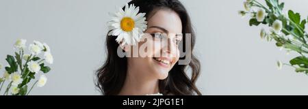 Jeune femme brune avec maquillage et camomille dans les cheveux debout près des fleurs isolées sur le gris, bannière Banque D'Images