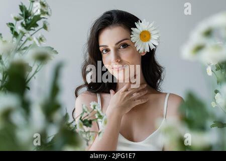 Portrait de femme souriante avec camomille dans les cheveux touchant le cou isolé sur gris Banque D'Images