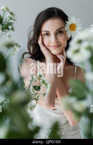 Jolie femme avec camomille dans les cheveux regardant la caméra près de fleurs floues isolées sur gris Banque D'Images