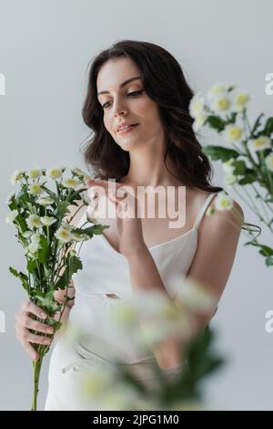 Jolie femme brune en blanc dessus touchant des fleurs isolées sur gris Banque D'Images