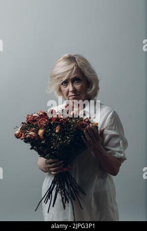 Triste femme âgée senior en chemise blanche tient avec une vieille fleur de rose mort sèche. Notion de vieillesse, de vieillissement et de mort. Banque D'Images