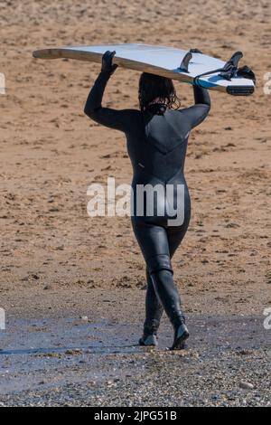 Vue arrière d'une surfeuse marchant et équilibrant une planche de surf sur sa tête après une séance de surf à Fistral Beach à Newquay, en Cornouailles, au Royaume-Uni. Banque D'Images