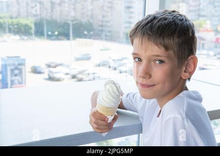 Un jeune garçon mignon mange une glace blanche tourbillonnante dans une tasse à gaufres, assis à une table près de la fenêtre dans un café, une copie de l'espace. Arrière-plan flou Banque D'Images