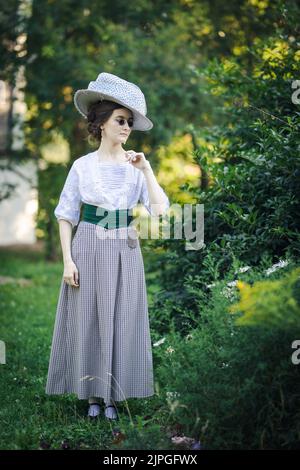 Portrait d'une jeune femme mince en costume de 1910s. Une dame dans un chapeau à la mode du début du 20th siècle marche dans un jardin Banque D'Images