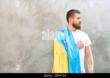 L'homme tient la main sur son coeur et son drapeau national bleu-jaune de l'Ukraine sur son épaule. Guy en T-shirt blanc et jeans. Jour ensoleillé. Jour de la Constitution et de l'indépendance de l'Ukraine. Mise au point sélective douce Banque D'Images