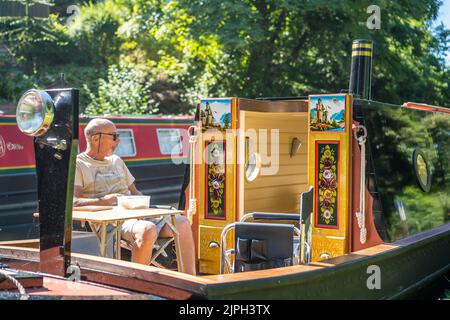 Le bateau à narrowboat britannique amarré au bord du canal de Worcestershire (portes ouvertes montrant la peinture traditionnelle des roses et des châteaux) tandis que l'homme est assis à profiter de la vague de chaleur. Banque D'Images