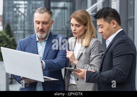 Groupe d'affaires diversifié composé de trois personnes discutant et discutant de la stratégie en dehors du bâtiment de bureau homme d'affaires et femme d'affaires regardant l'écran d'ordinateur portable en se tenant debout Banque D'Images