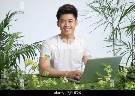 Portrait d'un beau et heureux jeune vietnamien travaillant sur ordinateur portable avec des plans verts autour Banque D'Images