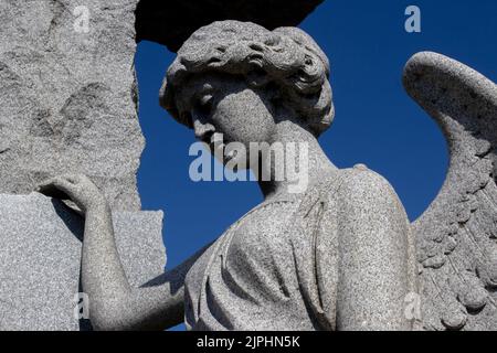 Un ange de pierre se tient avec sa main sur la croix et ses ailes se sont partiellement répandues avec un regard de tristesse et de perte contre le ciel bleu foncé Banque D'Images