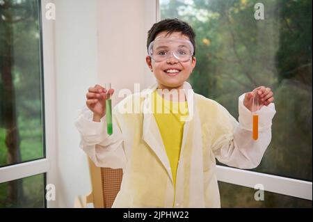 Gai petit chimiste scientifique, écolier en lunettes de sécurité et blouse de laboratoire tenant des tubes à essai, sur une classe de chimie Banque D'Images