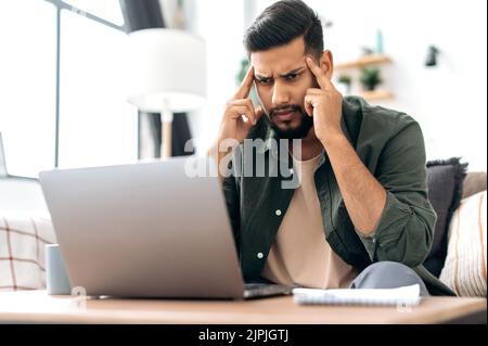 Tendus concentrés lassé arabe ou indien stylisé, indépendant, assis sur un canapé dans un salon, utilisant un ordinateur portable pour travailler ou étudier à distance, massant ses temples, se sentir surtravaillé, maux de tête Banque D'Images