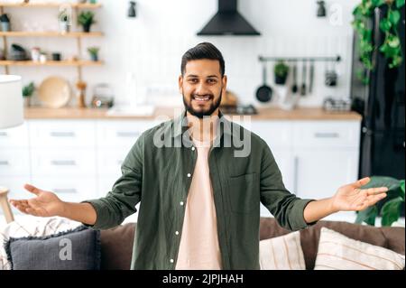 Joyeux joli garçon indien ou arabe moderne positif, dans des vêtements élégants et décontractés, se tenir à la maison dans le salon sur le fond de la cuisine, se répand ses bras sur le côté, regarde l'appareil photo, sourire Banque D'Images