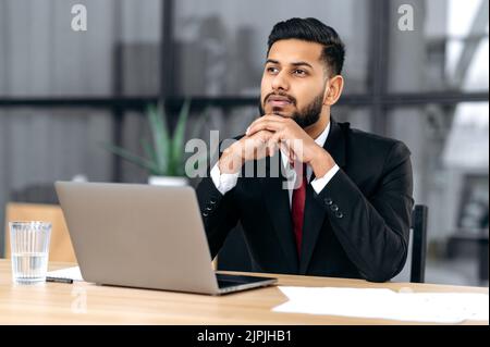 Un homme d'affaires influent et pensif, d'origine indienne ou arabe, assis à une table au bureau avec un ordinateur portable, regarde droit, réfléchit aux idées d'un nouveau projet, analyse les risques et les bénéfices Banque D'Images