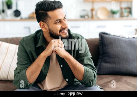 Beau positif confiant et attrayant indien ou arabe, dans des vêtements élégants et décontractés, assis à la maison dans le salon confortable sur le fond de la cuisine, regarde sur le côté, souriant Banque D'Images