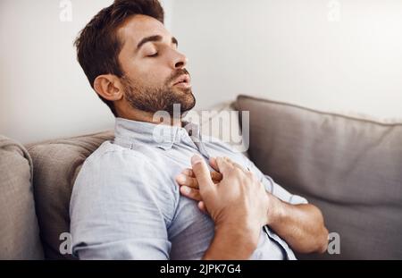 Espérons que cela n'est pas grave. Un jeune homme d'affaires charmant tenant sa poitrine en douleur tout en se relaxant sur un canapé à la maison. Banque D'Images