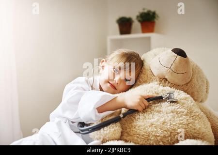 Teddy a le plus grand cœur que j'aie jamais vu. Une adorable petite fille vêtue comme un médecin et traitant son ours en peluche comme un patient. Banque D'Images