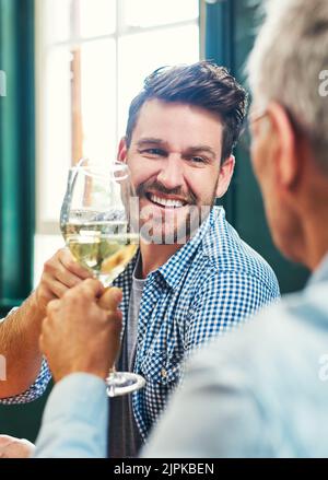 Nous devons le faire plus souvent. Un jeune homme joyeux et son père mûr ayant un toast festif avec des verres à vin à la maison. Banque D'Images