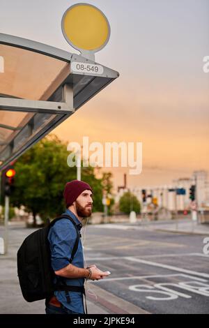 Ses trajets quotidiens commencent et se terminent toujours par de la musique. Un beau jeune homme qui écoute de la musique sur son téléphone portable en attendant à un arrêt de bus. Banque D'Images