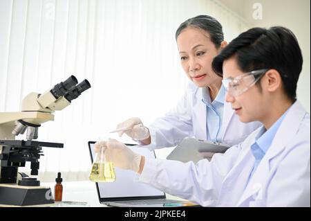 Une femme asiatique d'âge moyen professionnelle forme un scientifique et donne un conseil à un jeune scientifique pendant l'expérience chimique en laboratoire. Banque D'Images