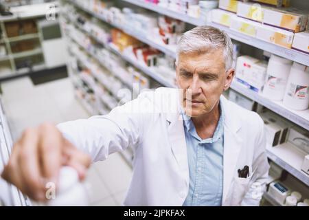 Ce n'est pas possible ici. Un pharmacien masculin mature et concentré qui replace une bouteille de médicament sur l'étagère d'une pharmacie. Banque D'Images