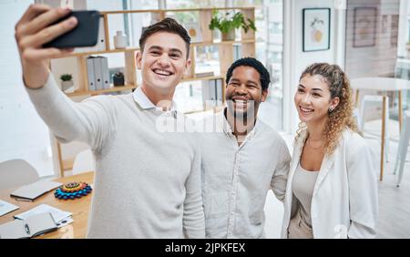 Groupe d'amis prenant un selfie, avec un téléphone et passant du temps ensemble dans un bureau moderne. Groupe multiracial de jeunes excités, heureux et riant Banque D'Images