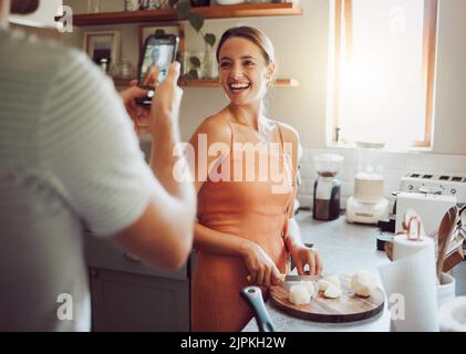 Femme cuisinant avec son mari prenant une photo dans la cuisine sur son téléphone portable à la maison. Beau chef, heureux et femme de couper des légumes pour cuisiner Banque D'Images