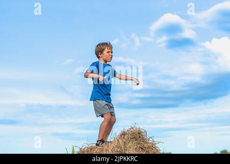 Garçon sourire jouer danse grimace spectacle de t-shirt bleu sur des balles de foin sec, ciel clair jour ensoleillé. Équilibrer la formation. Concept heureux Banque D'Images