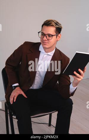 Élégant homme habillé en costume assis lisant un livre avec une expression heureuse. Homme sur fond blanc étudiant. Un enseignant donnant la classe avec satisfaction. Photo de haute qualité Banque D'Images