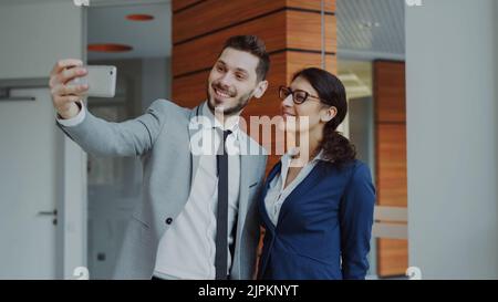 Un homme d'affaires heureux et sa collègue qui emont un selfie sur un appareil photo intelligent pour les médias sociaux dans un bureau moderne à l'intérieur Banque D'Images