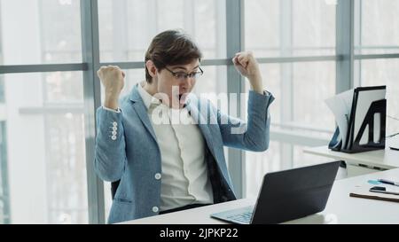 Jeune homme d'affaires utilisant un ordinateur portable recevant un bon message et devenir très excité et heureux assis dans le bureau moderne à l'intérieur Banque D'Images