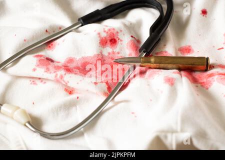 Un stéthoscope et une balle reposent sur une robe blanche avec des taches de sang rouges, des traces de sang sur les choses et une blessure à balle, assistance médicale Banque D'Images