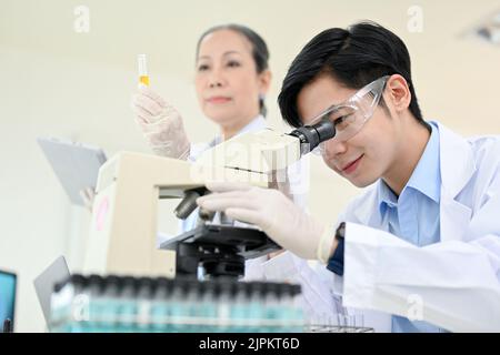 Un jeune scientifique asiatique expert ou un chimiste médical examinant un échantillon de virus sous microscope dans le laboratoire avec son superviseur féminin âgé. Banque D'Images