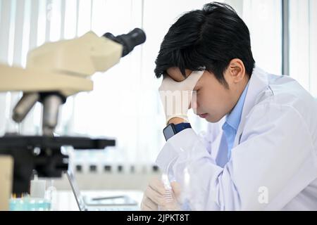 Un jeune homme asiatique stressé, un scientifique ou un technicien médical en blouse blanche, à son bureau, dans le laboratoire. Surtravaillé, contrarié, le travailleur de l'échec concep Banque D'Images