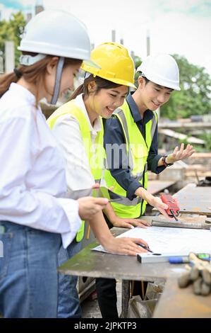 Équipe d'un ingénieur civil asiatique joyeux et professionnel du millénaire qui vérifie le plan d'ensemble sur le site de construction. Banque D'Images