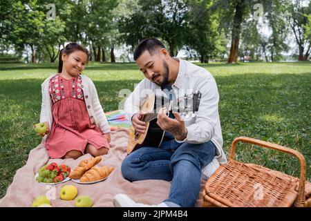 Homme asiatique jouant de la guitare acoustique près de la fille et de la nourriture dans le parc Banque D'Images