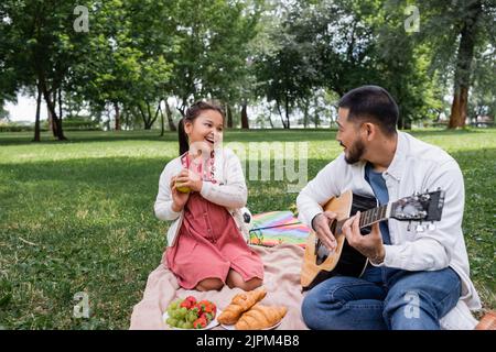 Joyeuse fille asiatique tenant la pomme près de père jouant de la guitare acoustique dans le parc Banque D'Images