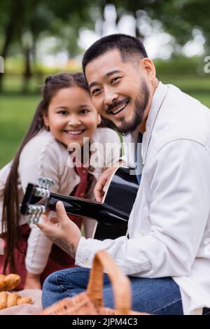 Homme asiatique positif jouant de la guitare acoustique et regardant la caméra près de la fille dans le parc Banque D'Images