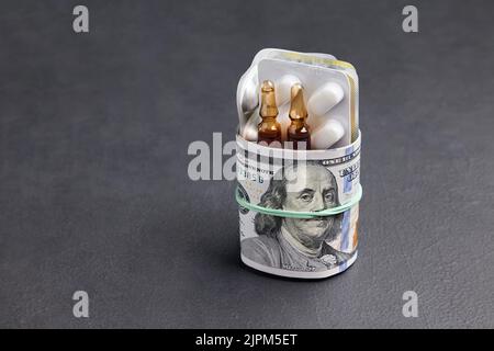 Les comprimés et les ampoules d'un blister sont emballés dans des billets de cent dollars, fixés avec une bande élastique sur un fond en béton noir. Banque D'Images