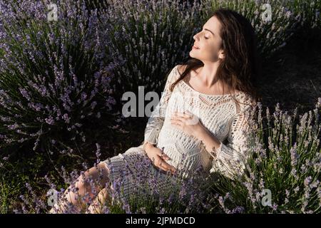une femme enceinte heureuse se touchant le ventre tout en étant assise dans un champ de lavande Banque D'Images