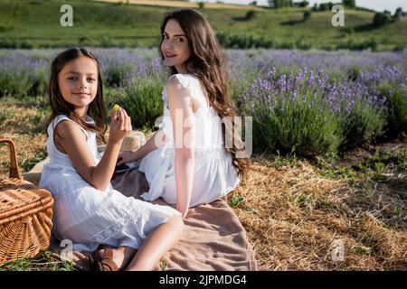 fille souriante tenant le raisin près de la mère sur le pique-nique dans le champ de floraison Banque D'Images