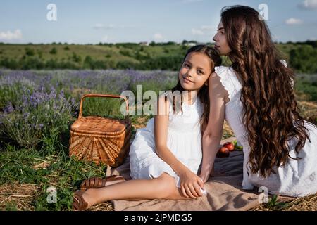 joyeuse fille regardant l'appareil photo près de brunette maman et panier de paille dans le pré de lavande Banque D'Images