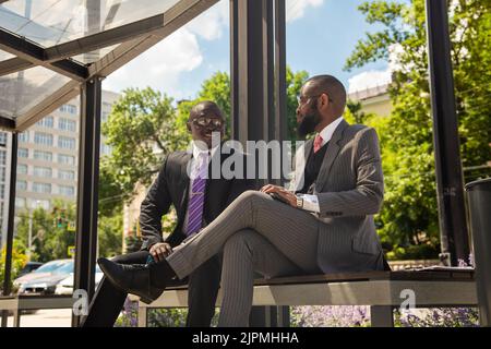 Deux hommes d'affaires à la peau sombre en costume communiquent. Les amis et les partenaires d'affaires sont assis sur un banc dans la ville sur fond de Banque D'Images